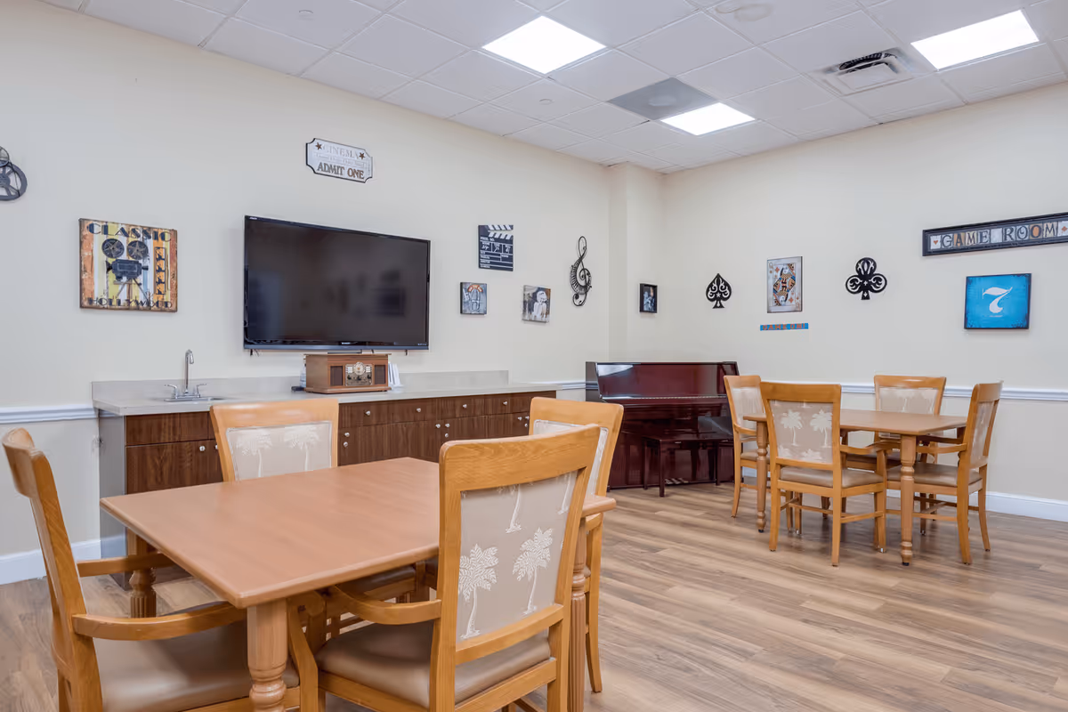 A game room with two wooden tables and chairs, a wall-mounted flat screen TV, a piano in the corner, and various decorative wall art including playing card symbols and a sign that says 'GAME ROOM'. The room has wood flooring and a light-colored ceiling with recessed lighting.