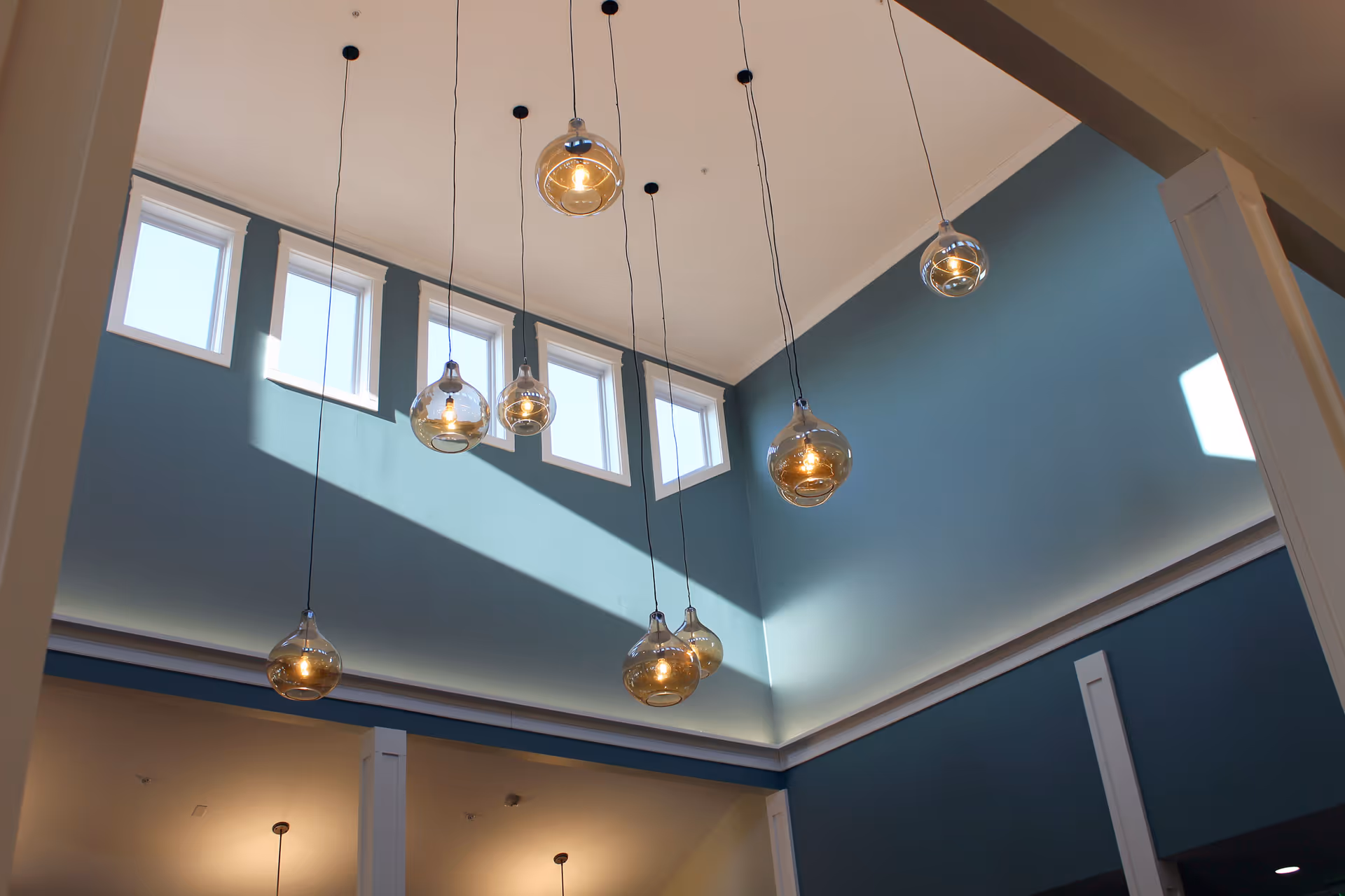 Interior view of a high ceiling room with multiple hanging pendant lights featuring round glass bulbs. The walls are painted blue with white trim, and there are five small rectangular windows near the ceiling letting in natural light.