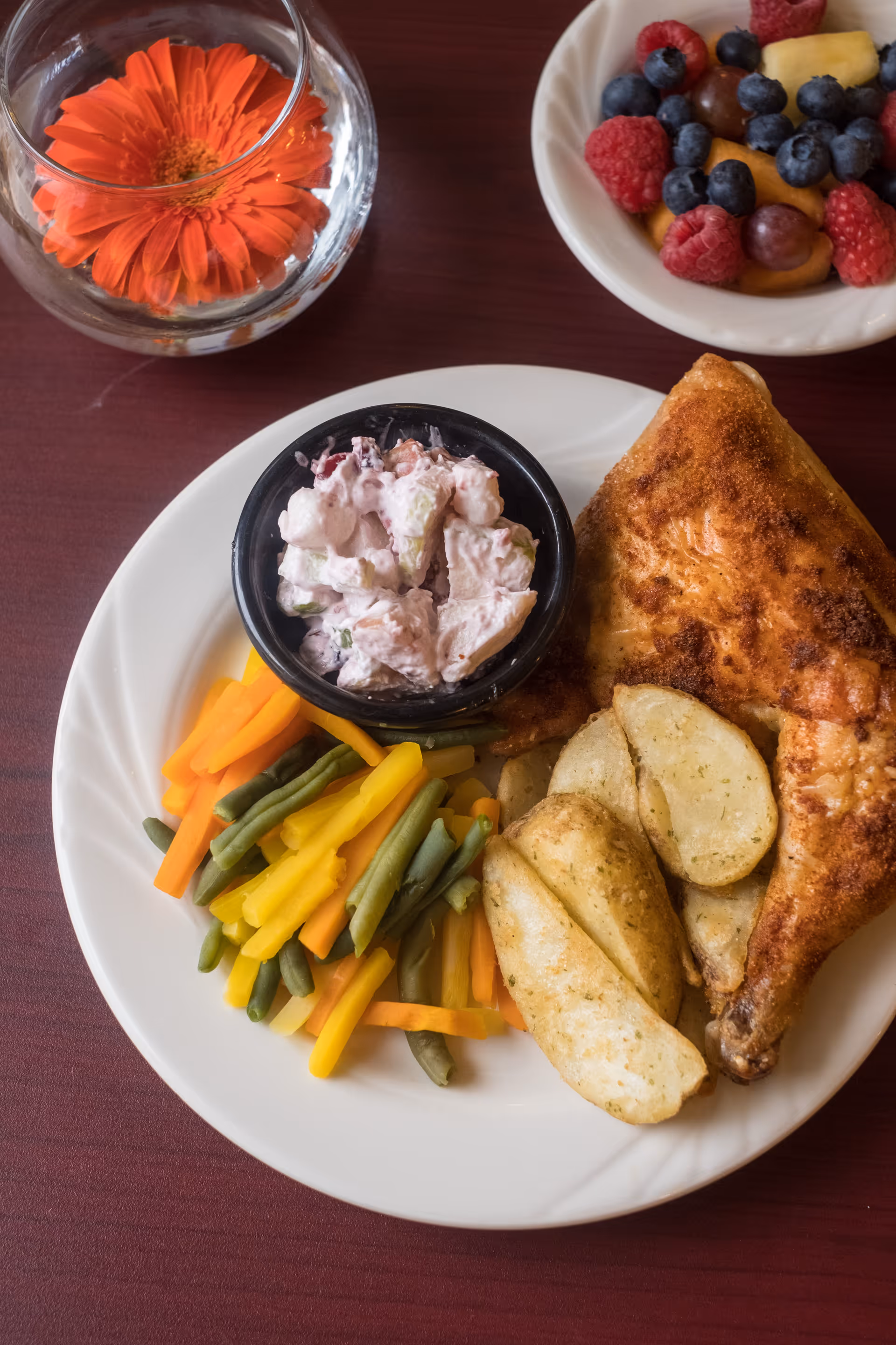 A plate of food featuring a seasoned baked chicken leg quarter, potato wedges, mixed steamed vegetables including carrots, green beans, and yellow beans, and a small bowl of creamy salad. Next to the plate is a bowl of mixed fresh fruit including raspberries, blueberries, grapes, and pineapple chunks. A glass vase with an orange flower is also visible on the table.