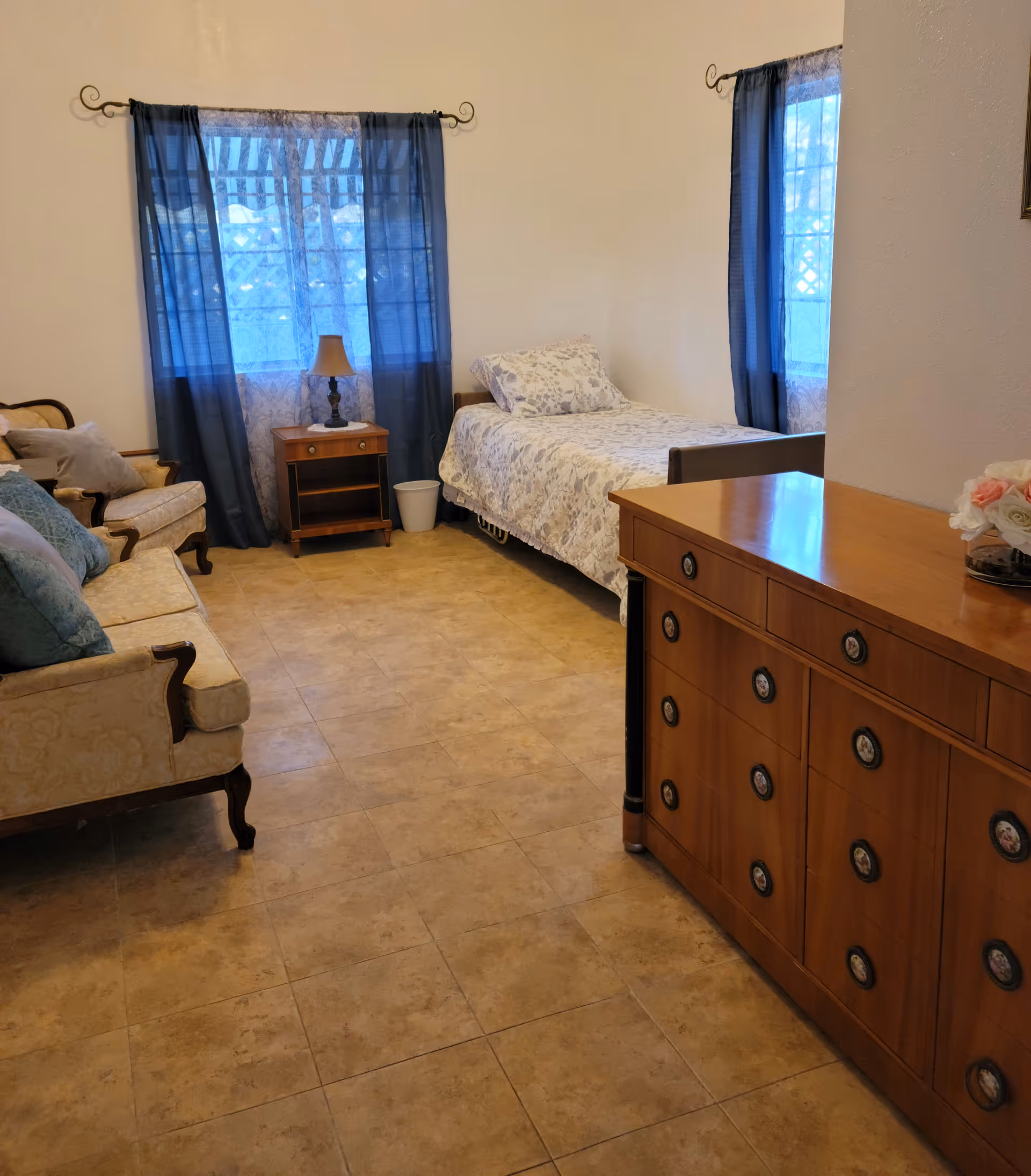 Furnished room with a single bed, sofa, wooden dresser and nightstand beneath blue curtained windows.