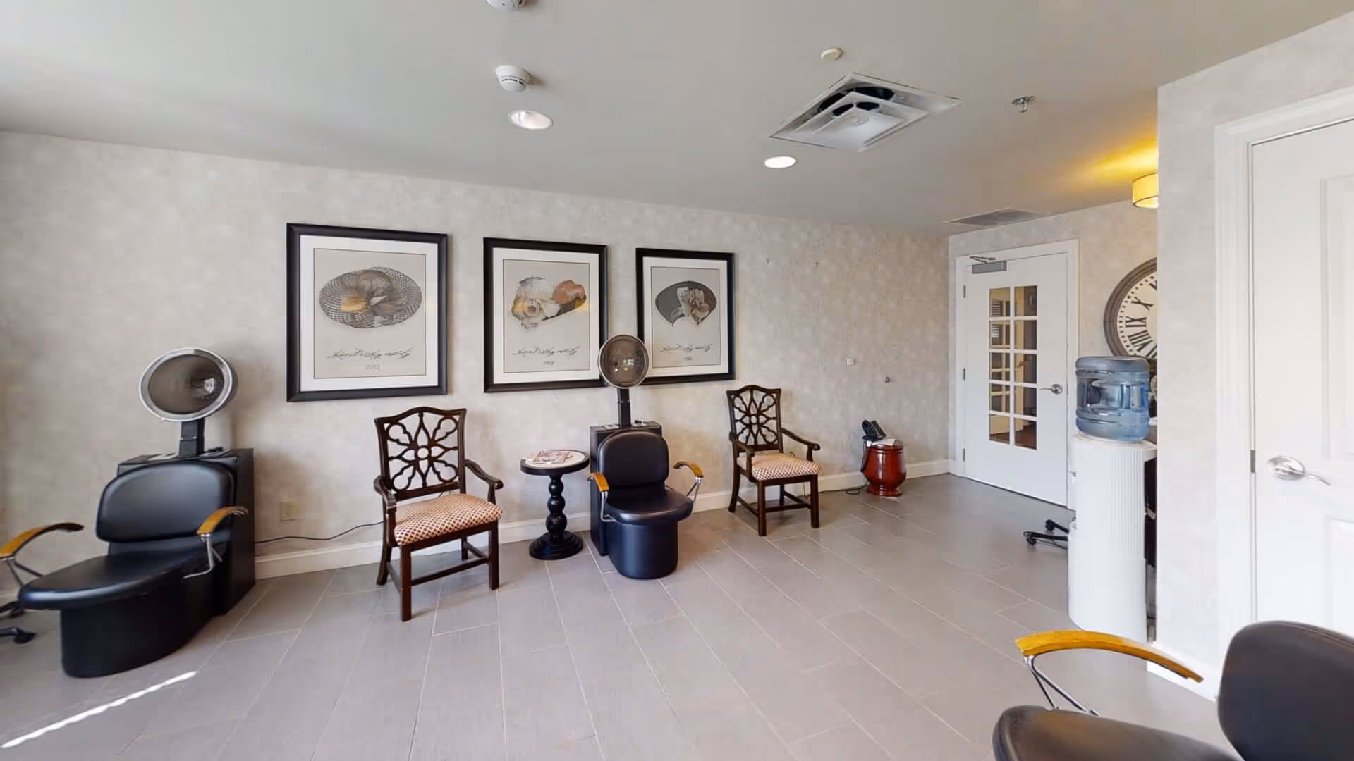 Interior view of a salon area in a senior living facility featuring two black salon chairs with hair dryers, two wooden chairs with patterned cushions, a small round table, framed artwork on the wall, a water cooler, and a large clock partially visible near a white door.