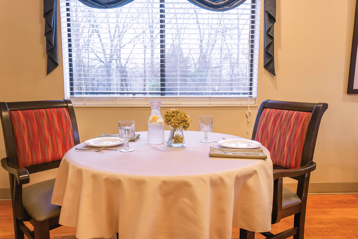 A small round dining table set for two with a white tablecloth, two plates, two glasses, silverware, a glass pitcher with lemon water, and a small vase with dried flowers. The table is positioned in front of a window with closed blinds and two wooden chairs with red and orange striped cushions.