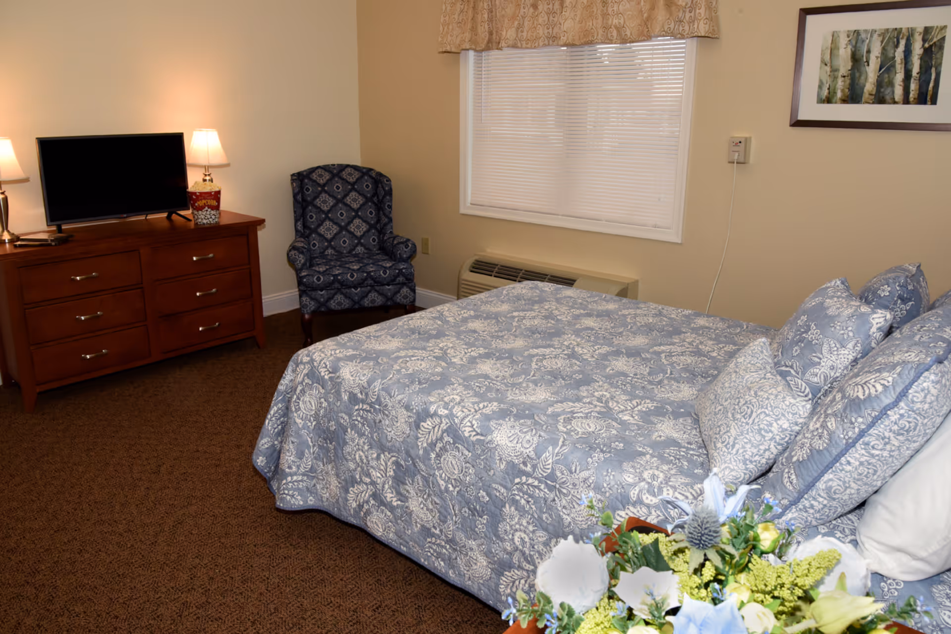 A furnished bedroom with a patterned blue bedspread, dresser with a TV, an upholstered chair, and a window.