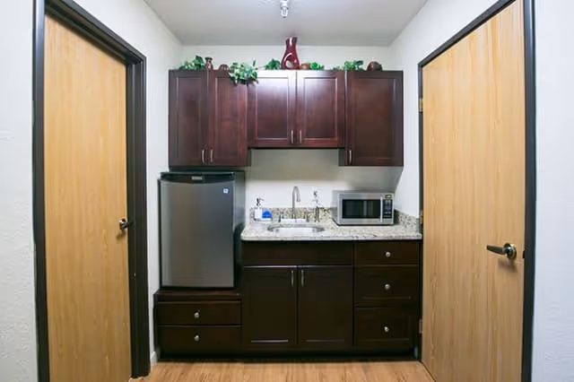 Small kitchenette with dark wood cabinets, granite countertop, mini fridge, sink, and microwave between two doors.