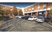 Exterior view of a multi-story brick building with several parked cars in front under a clear blue sky. Trees with autumn foliage are visible around the parking area.