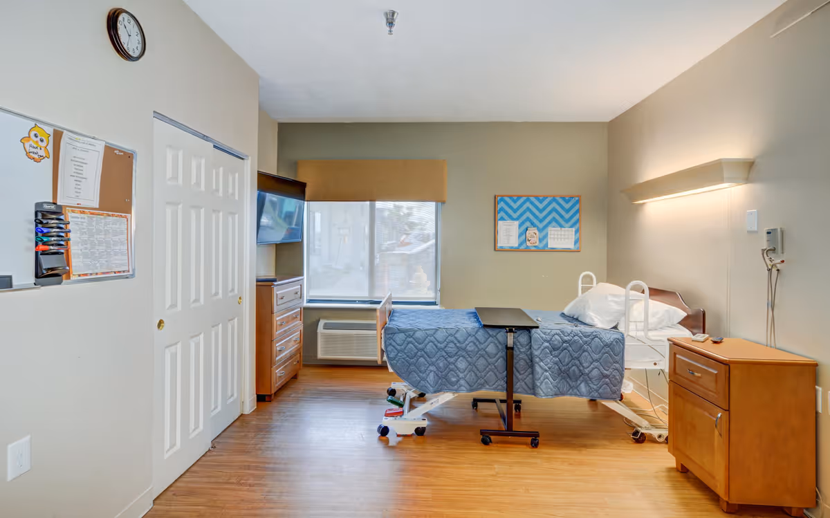 A healthcare facility room with a hospital bed covered in a blue quilt, a wooden nightstand with a lamp above it, a wooden dresser, a wall-mounted TV, a window with a brown valance, a clock on the wall, and a bulletin board with papers pinned on it.