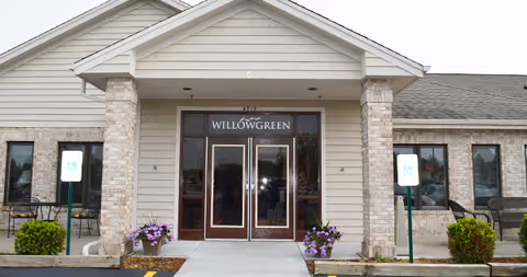 Front entrance of a single-story assisted living building with double glass doors under a 'WILLOWGREEN' sign, flanked by brick columns, potted flowers, and parking signs.