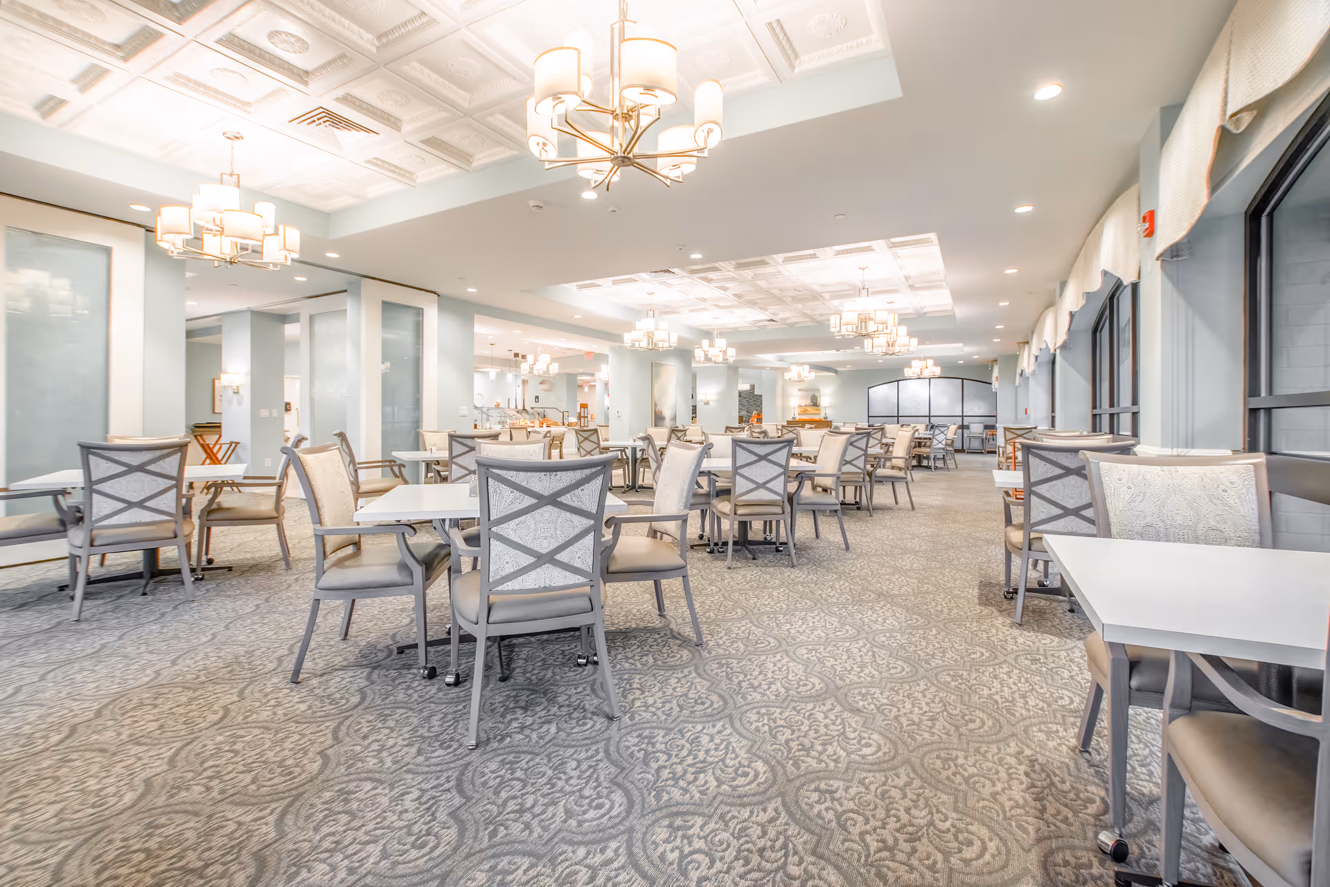 A spacious and well-lit dining room with multiple tables and chairs arranged neatly. The room features elegant chandeliers hanging from a decorative ceiling, large windows with curtains on one side, and a patterned carpet covering the floor. The overall ambiance is bright and inviting.