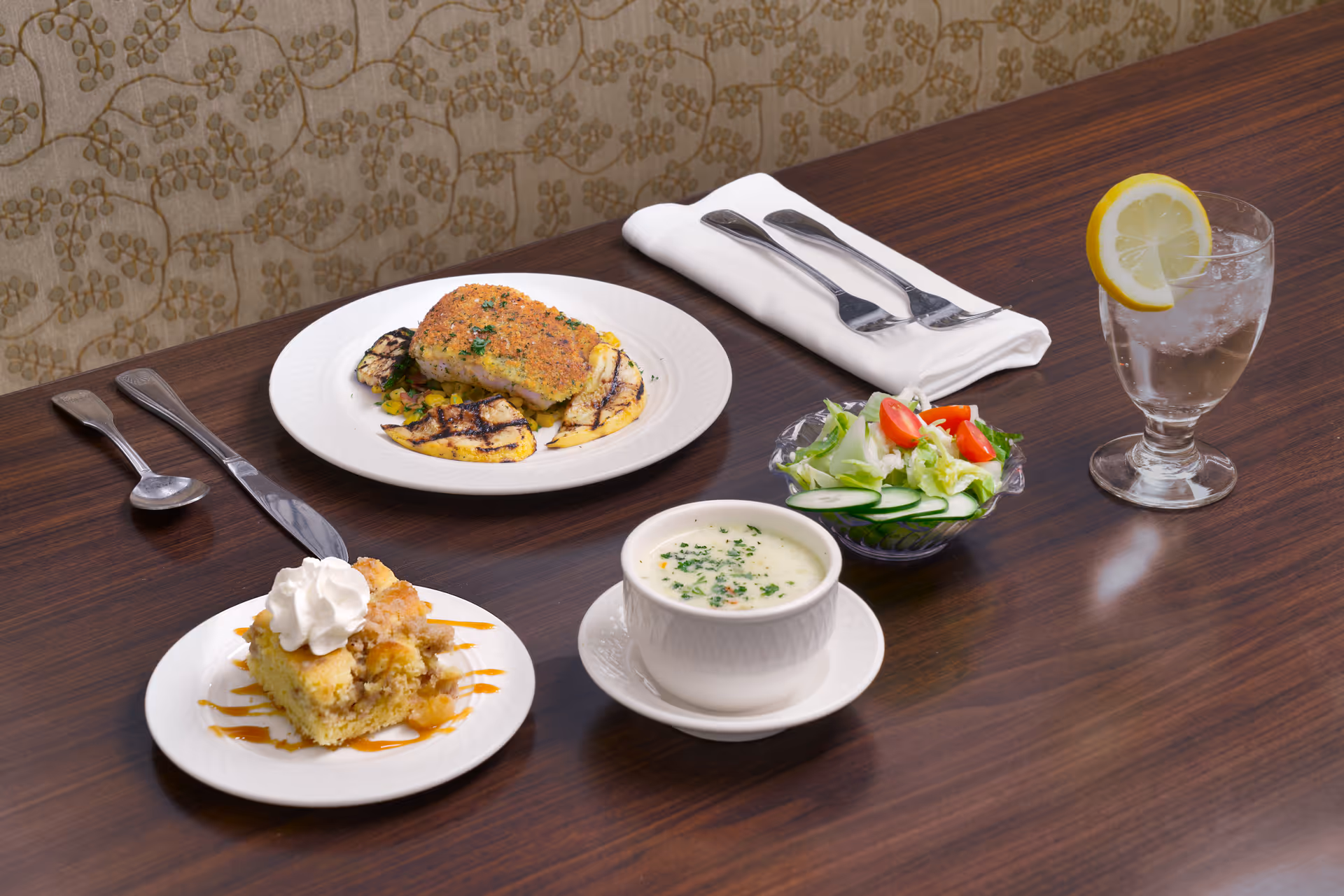 A dining table with plated meal including a main course, bowl of soup, small salad, dessert, utensils and a glass of water with a lemon slice.