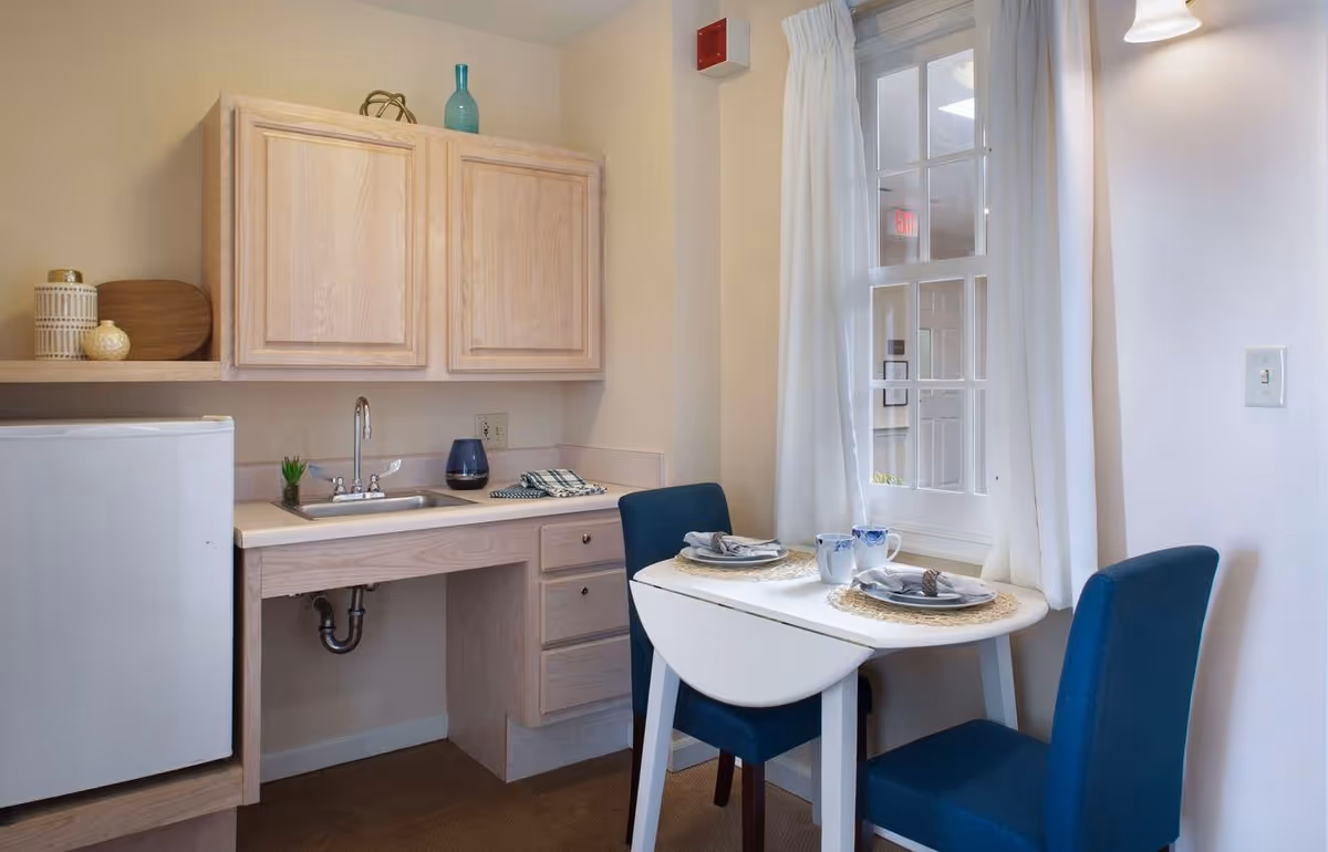 Small kitchenette and dining nook with a sink, light wood cabinets, mini-fridge and a two-seat table by a window.