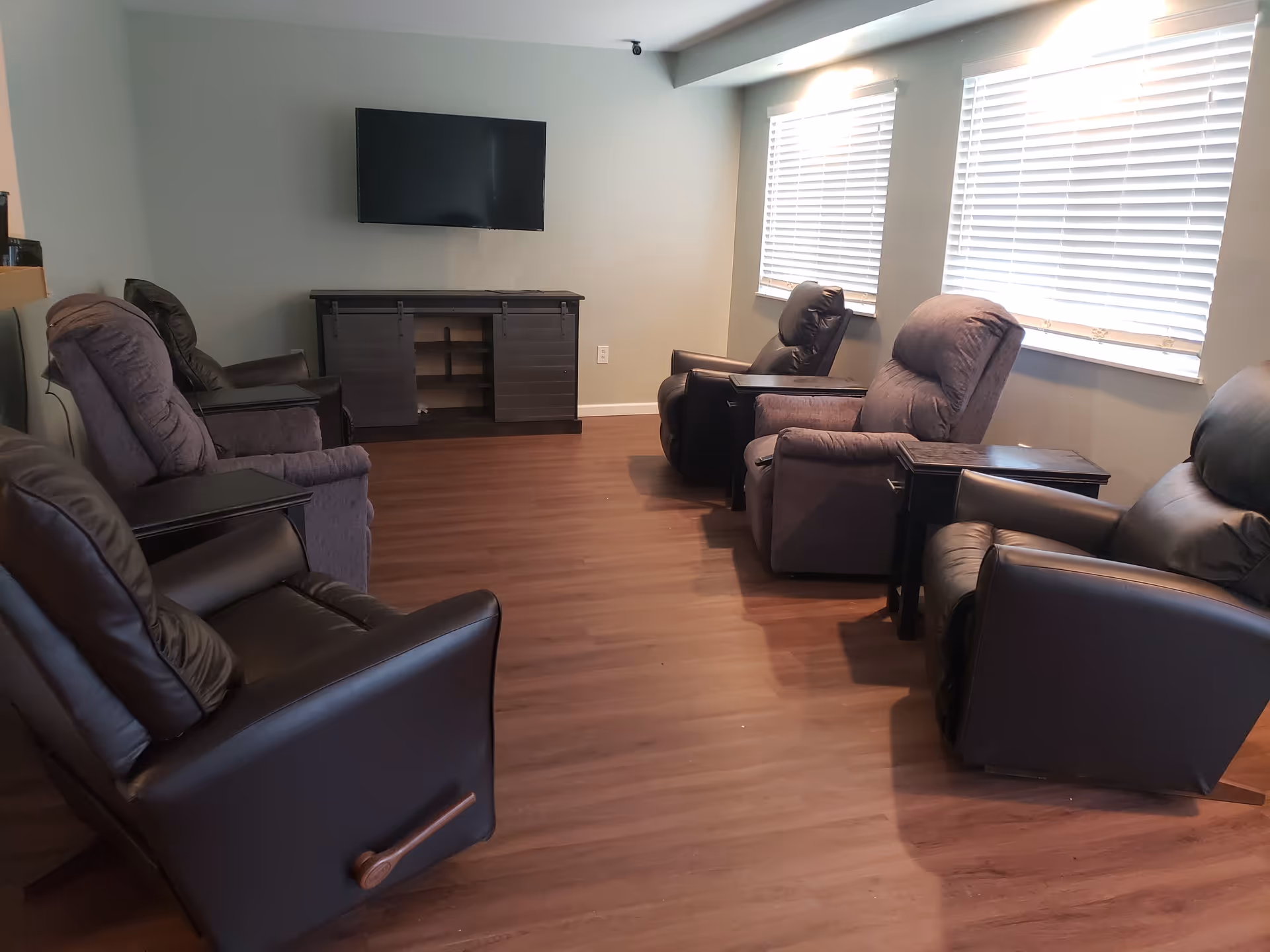 A cozy living room with six recliner chairs arranged in two rows facing a wall-mounted flat screen TV above a dark wooden cabinet. The room has wooden flooring and two windows with white blinds letting in natural light.