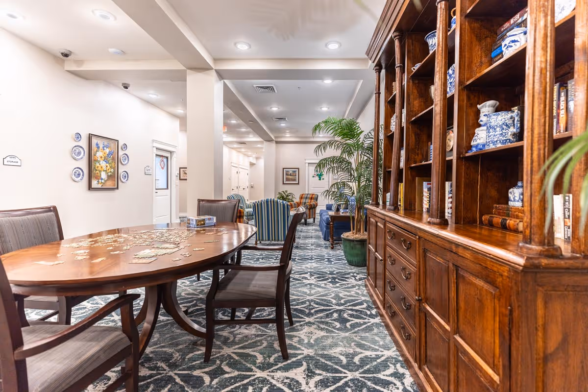 A well-lit common area in an assisted living facility featuring a wooden table with puzzle pieces on it and chairs around it. To the right is a large wooden bookshelf filled with books and decorative items. In the background, there are several upholstered armchairs and a potted plant, with white walls and ceiling lights illuminating the space.