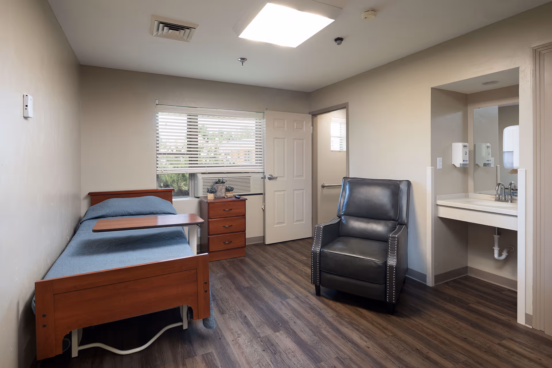 A simple, clean room in a healthcare facility with a single wooden bed covered with a blue blanket, a small wooden dresser with a plant and remote on top, a black leather armchair, a sink with a mirror and soap dispensers, and a window with blinds partially open letting in natural light.