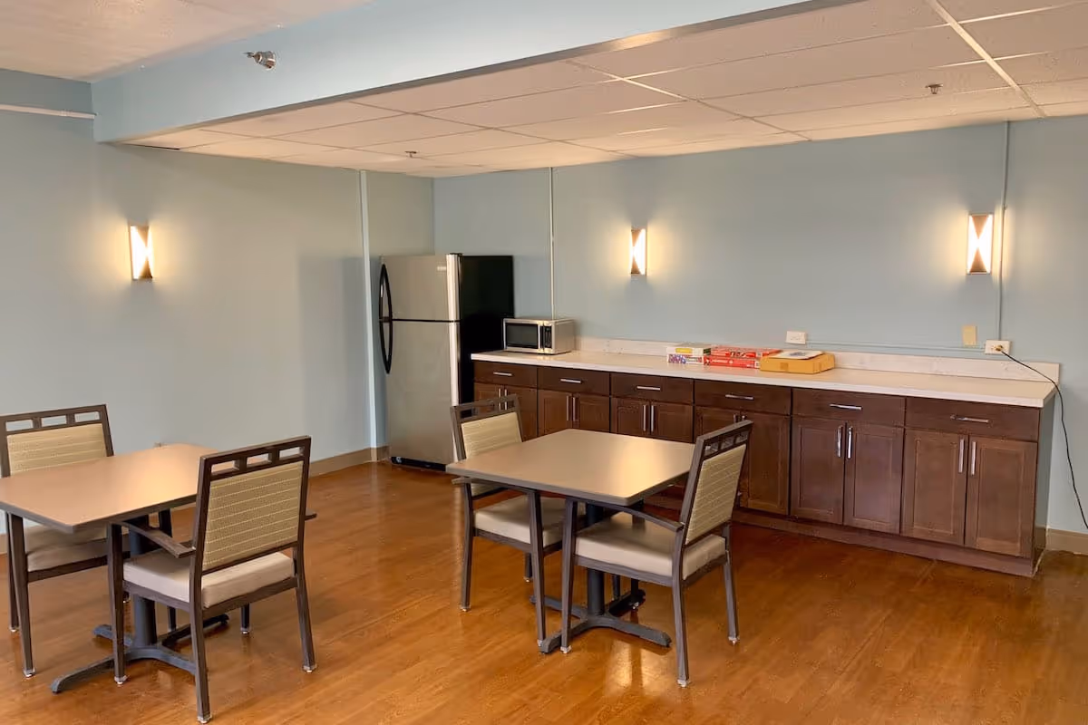 A small dining area with two square tables, each surrounded by four chairs. Against the wall is a countertop with dark wooden cabinets below, a microwave, and a stainless steel refrigerator. The walls are painted light blue with three modern wall sconces providing light. The floor is wooden.