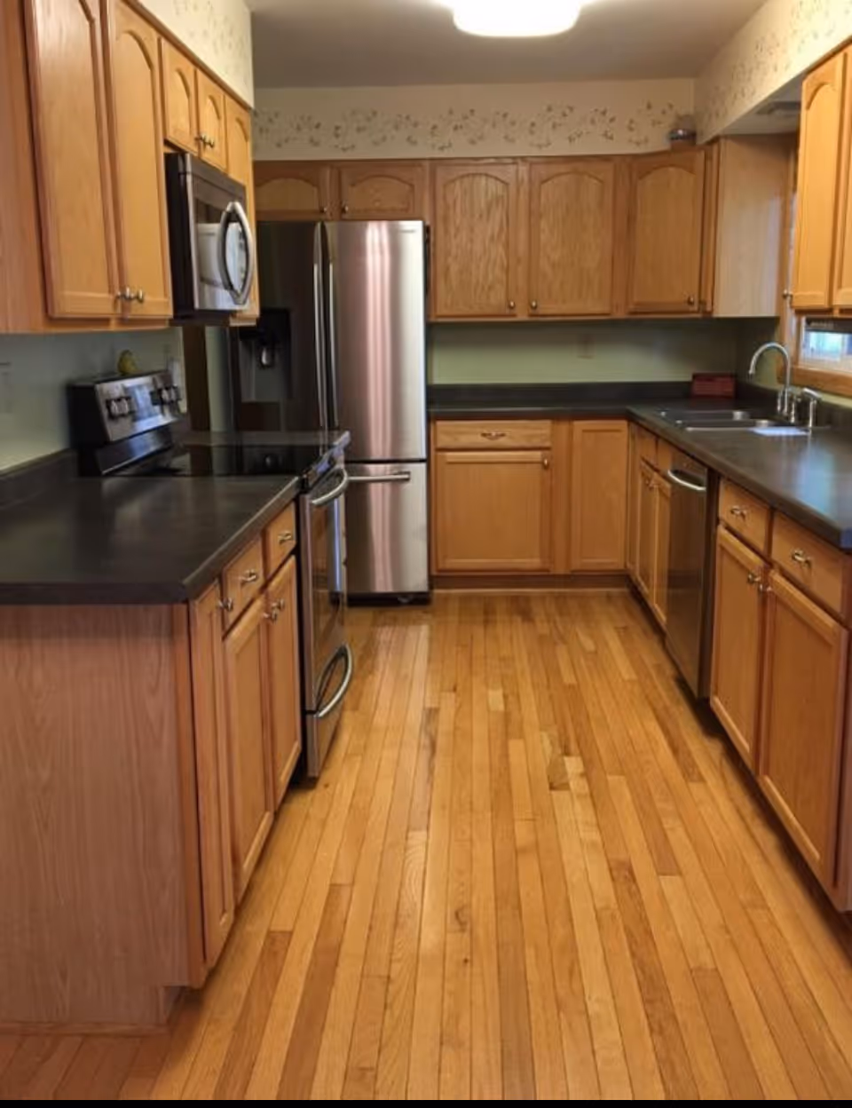 A kitchen with wooden cabinets and drawers, black countertops, a stainless steel refrigerator, stove, microwave, dishwasher, and a double sink. The floor is made of light-colored hardwood, and there is a window above the sink.