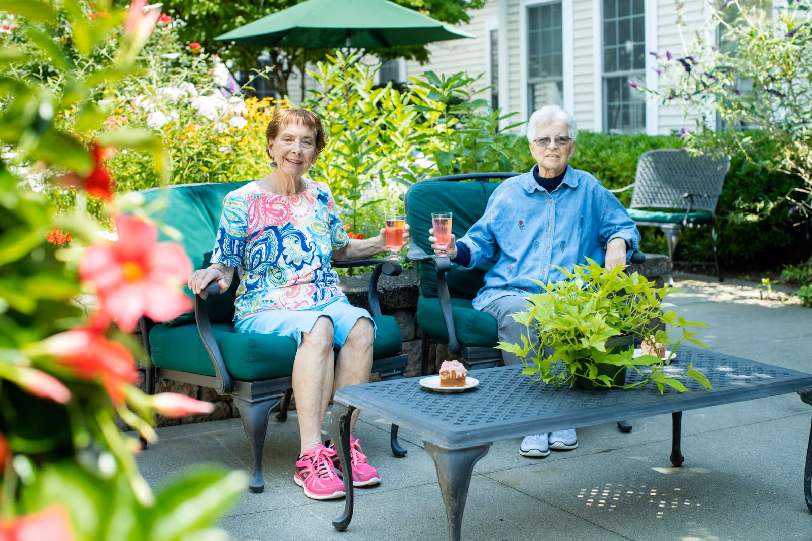 Two elderly women sitting outdoors on green cushioned chairs, holding glasses with a pink beverage. They are surrounded by lush greenery and flowers, with a table in front of them holding a potted plant and a plate with a piece of cake. A building with windows is visible in the background.