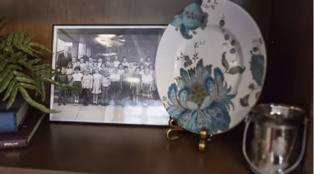 A wooden shelf displaying a framed black and white photograph of a group of children and an adult, a decorative plate with a blue and green floral pattern on a stand, a small silver cup, and a green leafy plant on the left side.