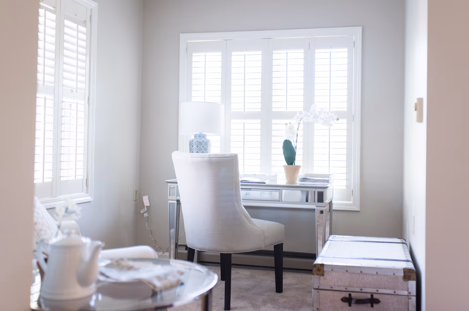 A bright room with a white upholstered chair in front of a mirrored desk, a table lamp with a blue patterned base, and a potted white orchid on the desk. The room has large windows with white plantation shutters, a white trunk with metal accents, and a glass table with a white teapot and a book.