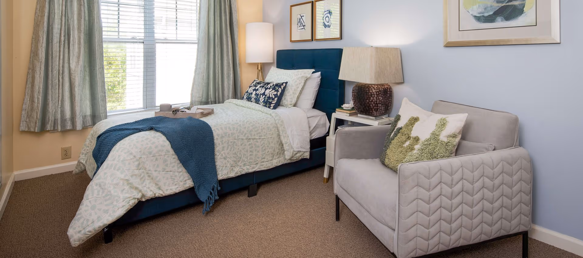 Bright single bedroom with a bed, bedside table and lamp, an upholstered armchair, and a window with curtains.
