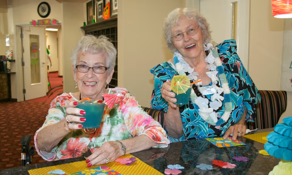 Two elderly women sitting at a table indoors, smiling and holding blue tropical drinks garnished with small umbrellas and straws. One woman is wearing glasses and a floral top, while the other wears a blue patterned dress with a white lei. The background shows a hallway and some wall decorations.