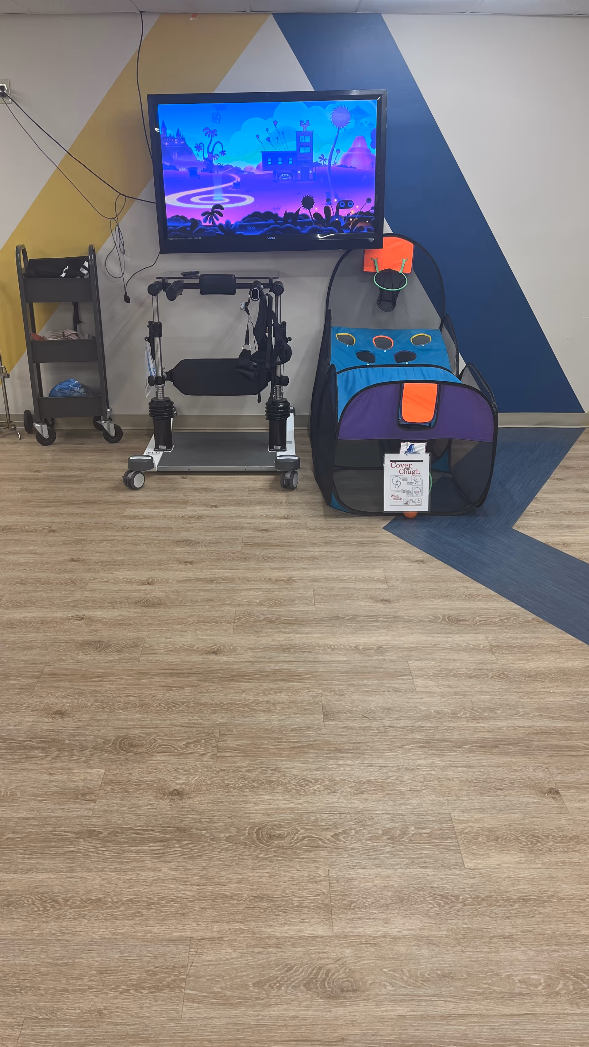 A room with a mounted flat-screen TV displaying a colorful animated scene. Below the TV, there is a black mobility aid device with wheels and a padded seat. To the right, there is a colorful play tent with a basketball hoop and holes for tossing balls. On the left side, there is a small black cart with shelves holding various items. The wall behind has a geometric pattern with yellow, white, and blue diagonal stripes. The floor is light wood with a blue accent area.