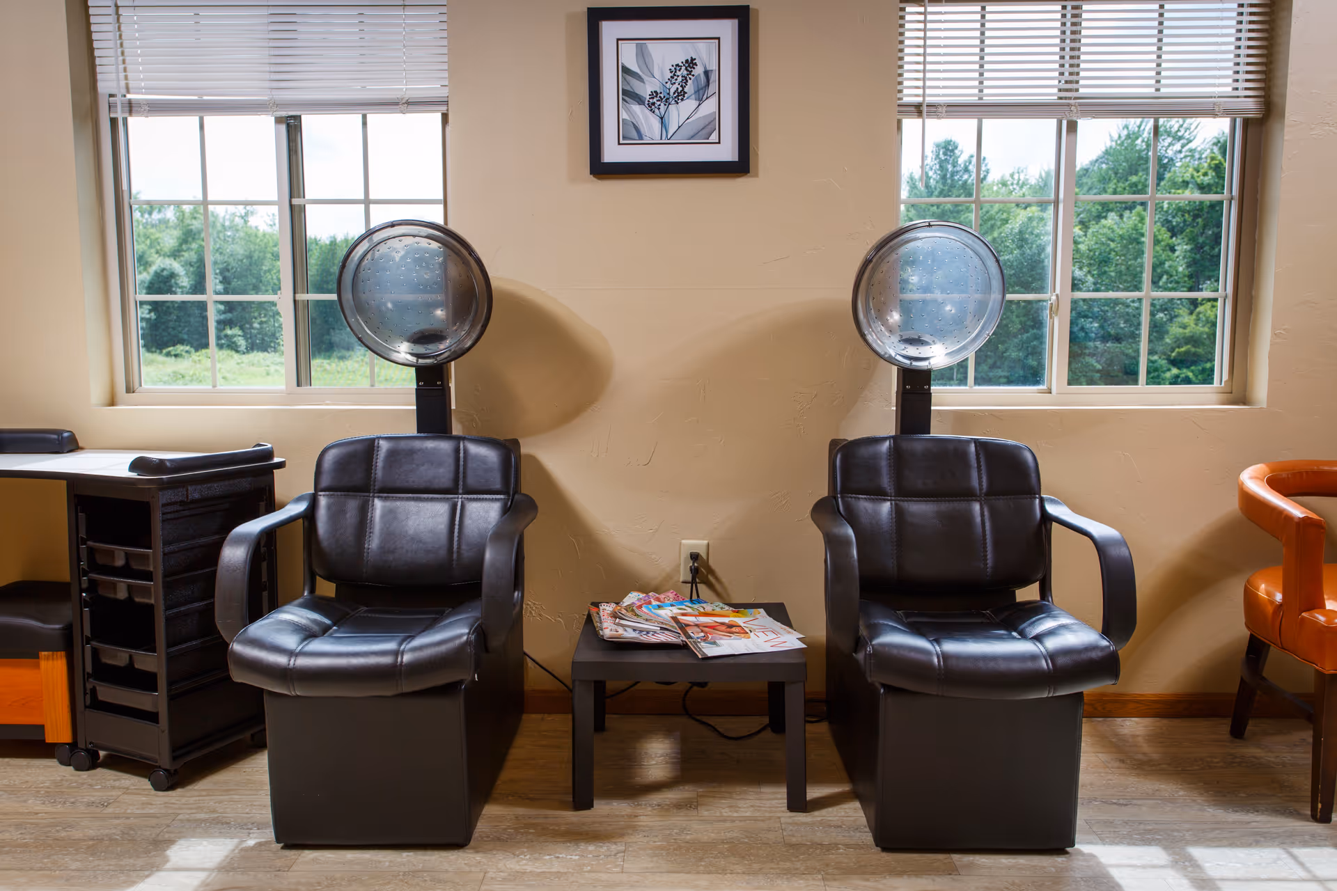 Two black salon chairs with overhead hair dryers sit in front of windows with a small table of magazines between them.
