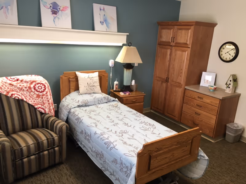 A cozy bedroom in a senior living facility with a single wooden bed covered with a light blue floral bedspread and a pillow that says 'Love you Mom'. Next to the bed is a wooden nightstand with a lamp, tissues, and a sanitizer bottle. There is a striped armchair with a red and white patterned blanket draped over it. On the wall above the bed are three watercolor paintings of animals. Across from the bed is a wooden wardrobe and a dresser with a clock on the wall above it and a small trash can nearby.
