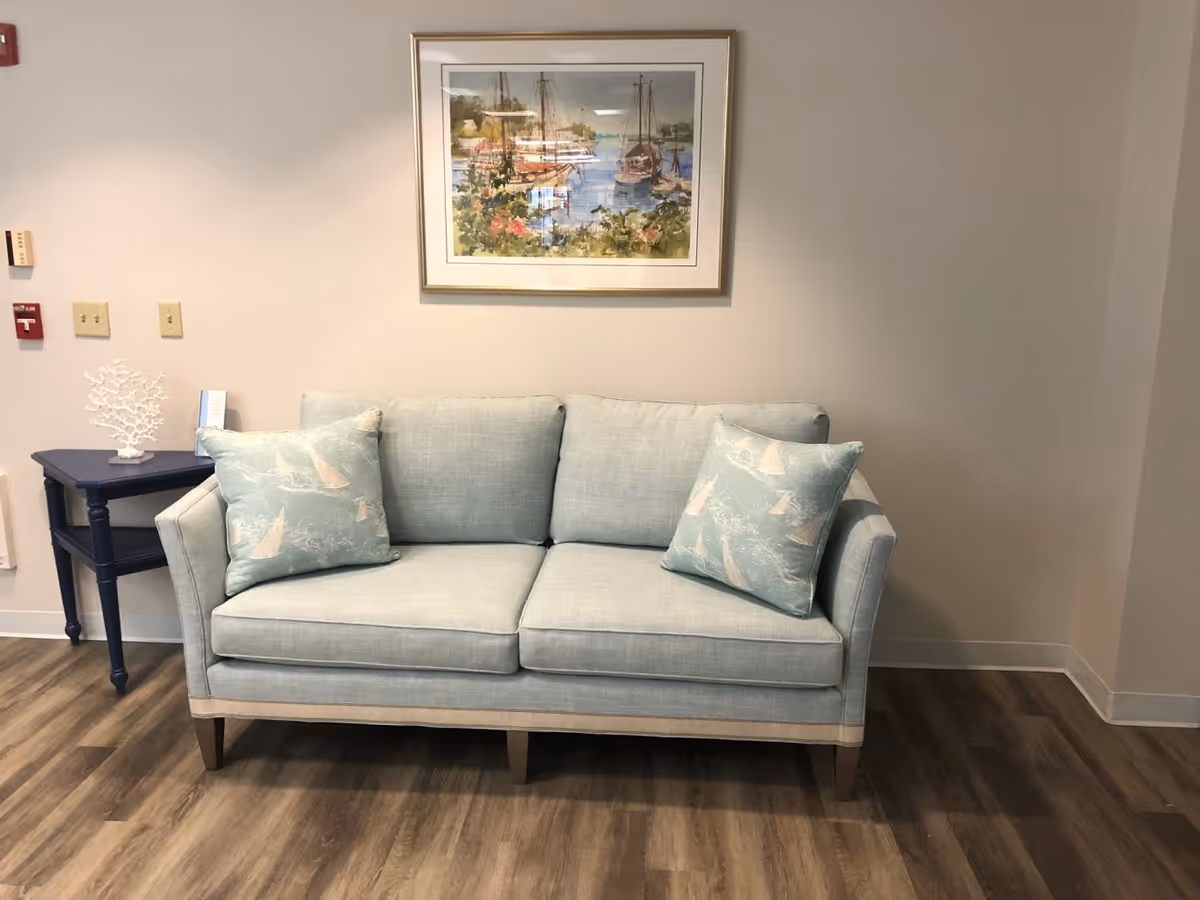 A light blue upholstered sofa with two matching cushions featuring sailboat patterns is placed against a beige wall. Above the sofa hangs a framed painting of boats docked at a marina with greenery and flowers in the foreground. To the left of the sofa is a small dark blue side table with a white coral decoration and some papers. The floor is wood with a medium brown finish.