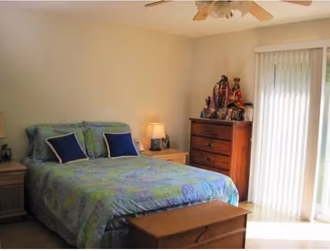 A bedroom with a bed covered in a blue and green patterned bedspread with two blue pillows. There are two wooden nightstands with lamps on either side of the bed. A wooden chest of drawers with decorative items and framed photos is against the wall near sliding glass doors with vertical blinds, allowing natural light into the room. A ceiling fan is visible above.