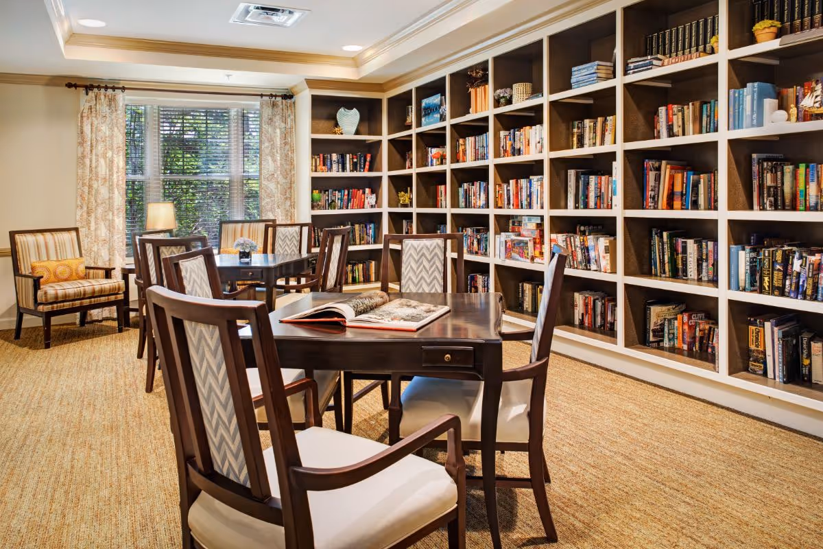 A cozy library room with built-in bookshelves filled with books along one wall. There are two dark wooden tables with cushioned chairs around them, and an open book lies on one table. A window with patterned curtains lets in natural light, and a striped armchair with a decorative pillow is placed near the window. The room has a warm, inviting atmosphere with beige carpeting and soft lighting.
