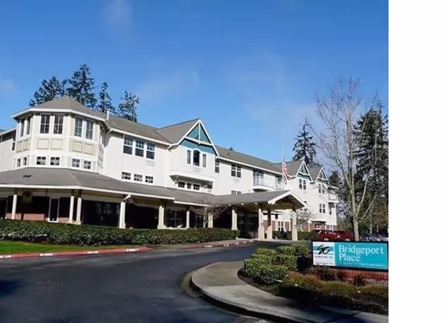 Exterior view of Bridgeport Place Assisted Living facility, showing a large multi-story building with a covered entrance, surrounded by trees and landscaping under a clear blue sky.