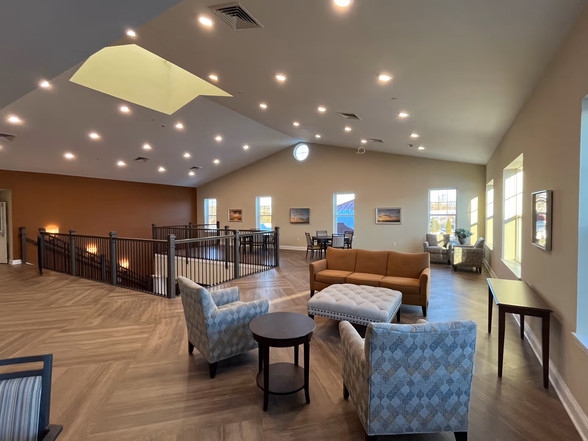 Spacious, well-lit common lounge with sofas, armchairs, tables, and large windows under a vaulted ceiling.