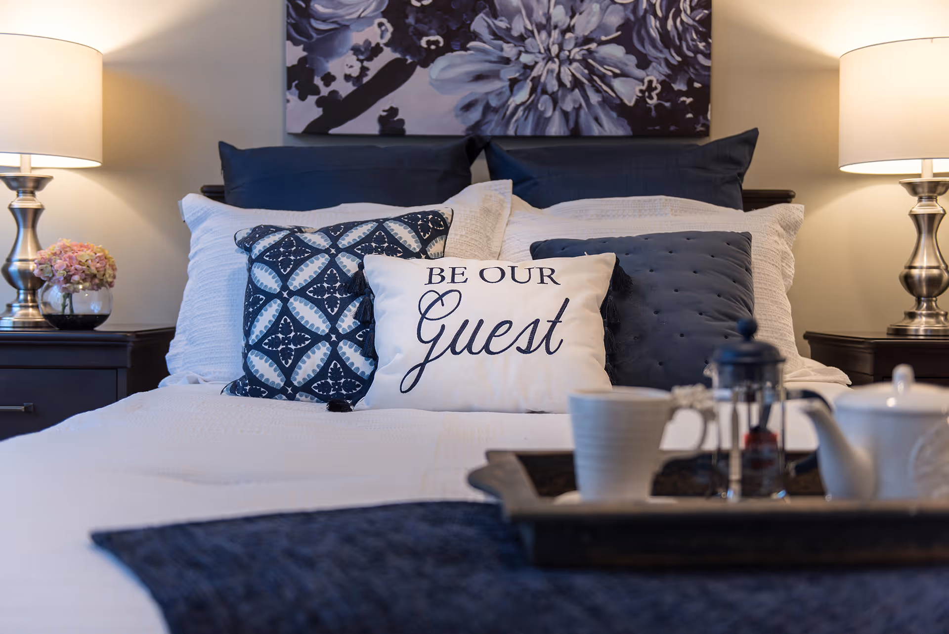 A neatly made bed in a bedroom with decorative pillows including a 'Be Our Guest' cushion, bedside lamps, and a breakfast tray.