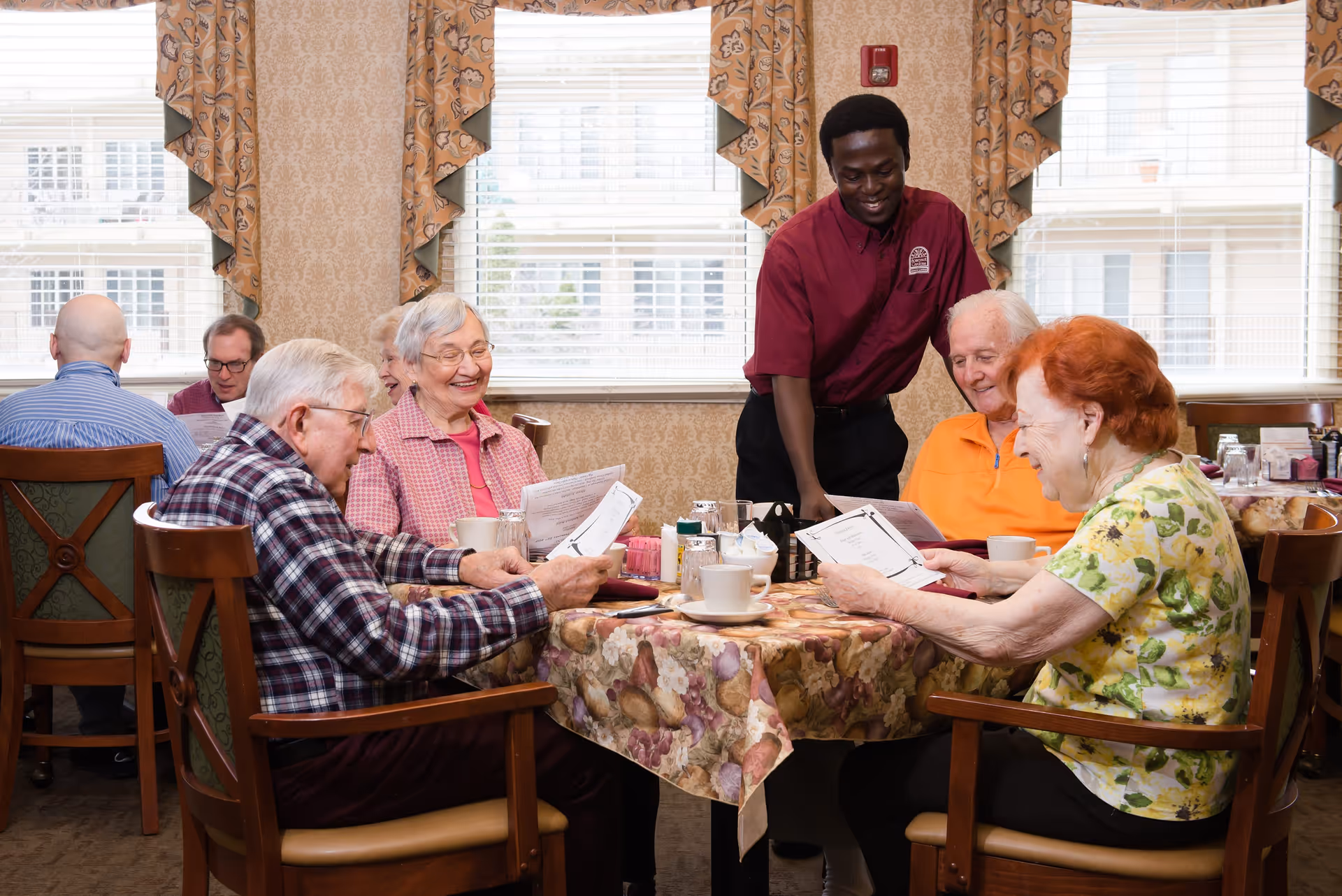 A group of elderly people sitting around a table in a dining room, smiling and looking at menus, while a staff member in a maroon shirt stands and interacts with them. The room has large windows with patterned curtains and a floral tablecloth on the table.