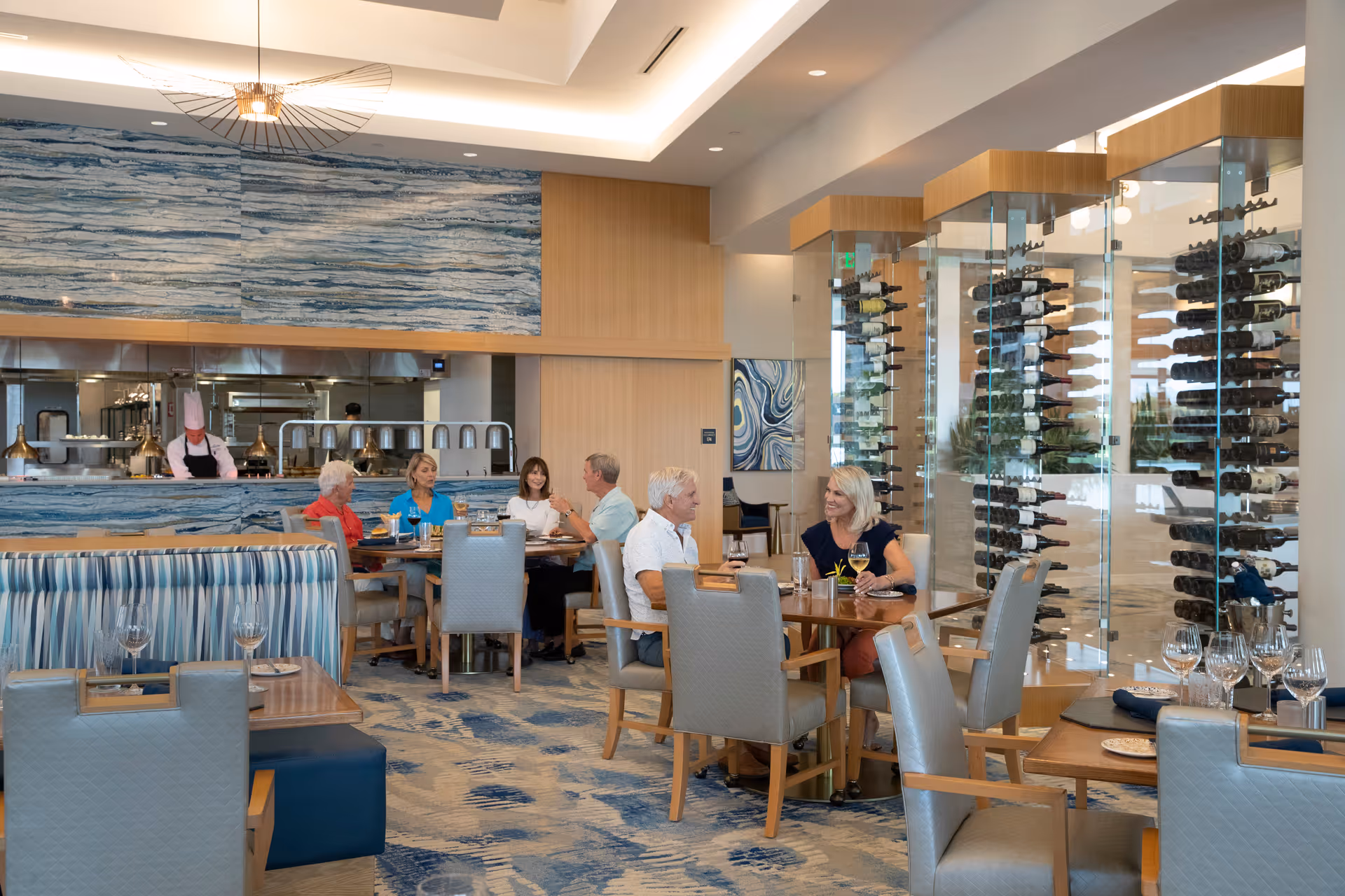 A modern dining room with several people seated at tables enjoying a meal. The room features a blue and beige color scheme with patterned carpet and wall art. There is a glass-enclosed wine display on the right side and an open kitchen area with a chef in the background.