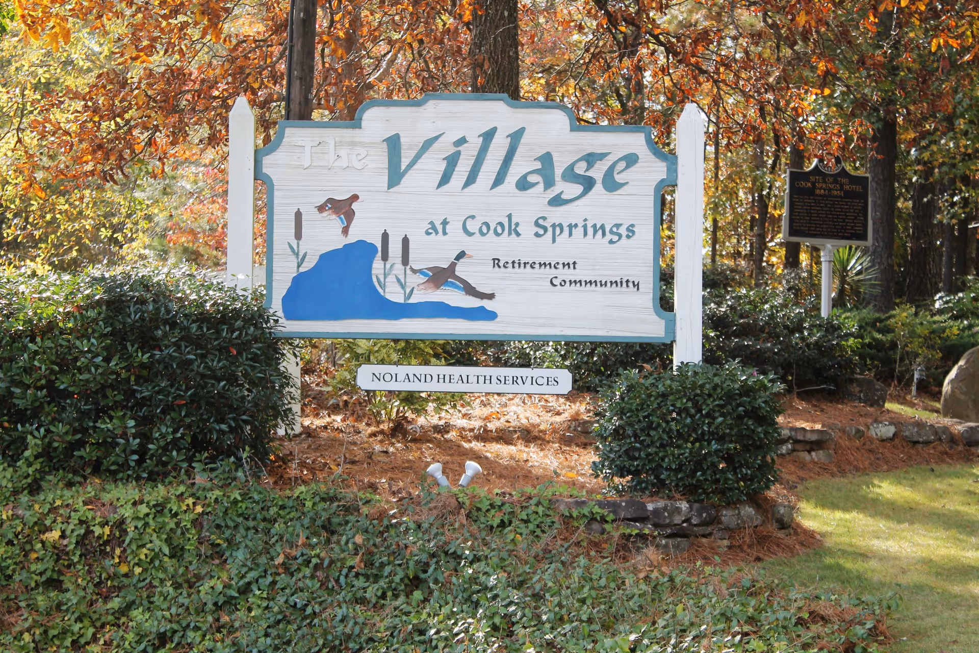 Wooden entrance sign reading "The Village at Cook Springs Retirement Community" surrounded by shrubs and trees.