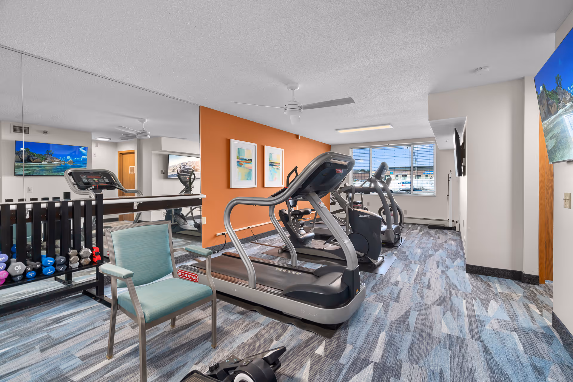 Fitness room with treadmills and other cardio equipment, dumbbells, seating, and a mirrored wall.