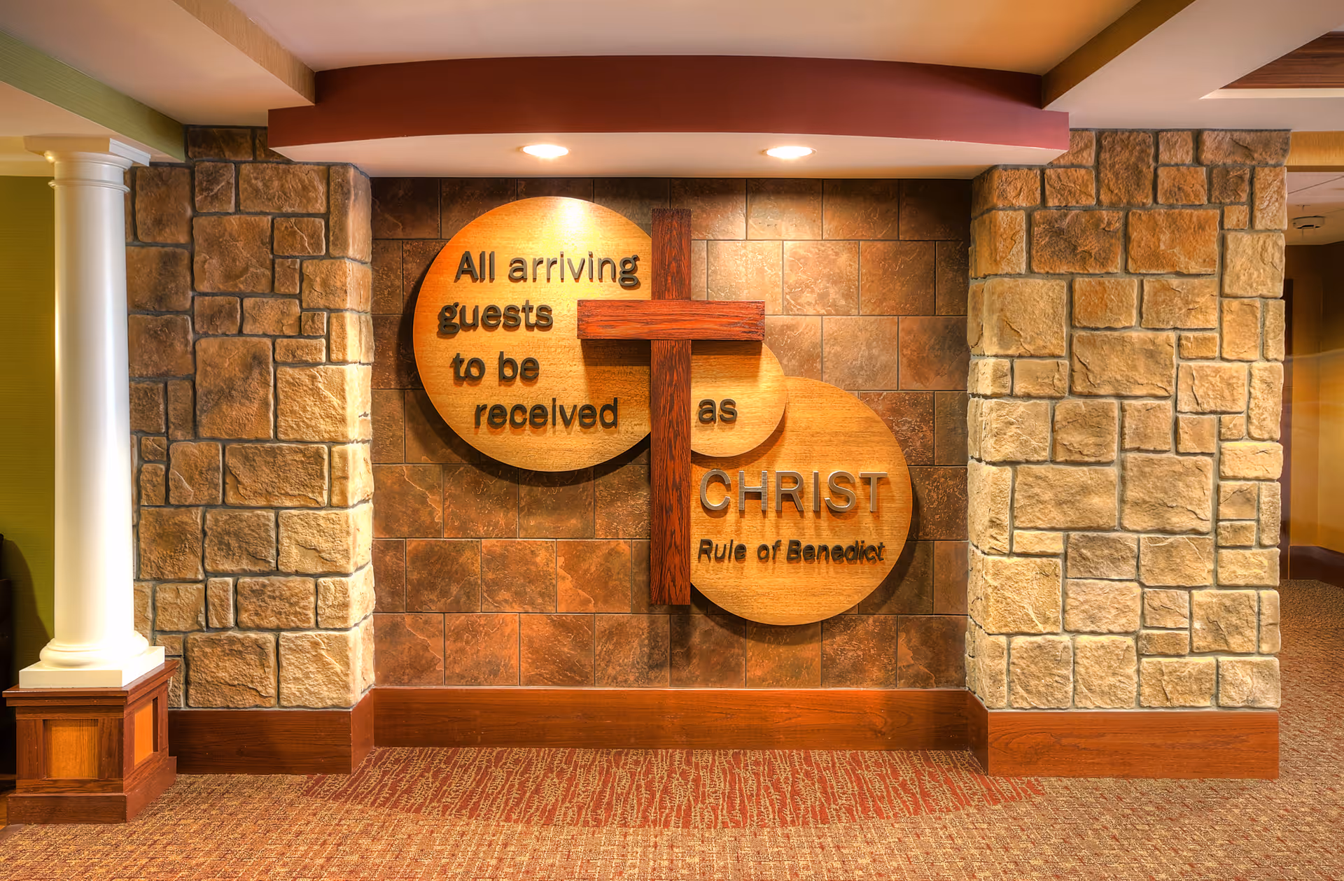 Interior wall with stone pillars and a wooden cross mounted on two overlapping wooden circles. The circles have the text: 'All arriving guests to be received as CHRIST Rule of Benedict'. The wall is tiled with brown tiles and the floor is carpeted in a red pattern.