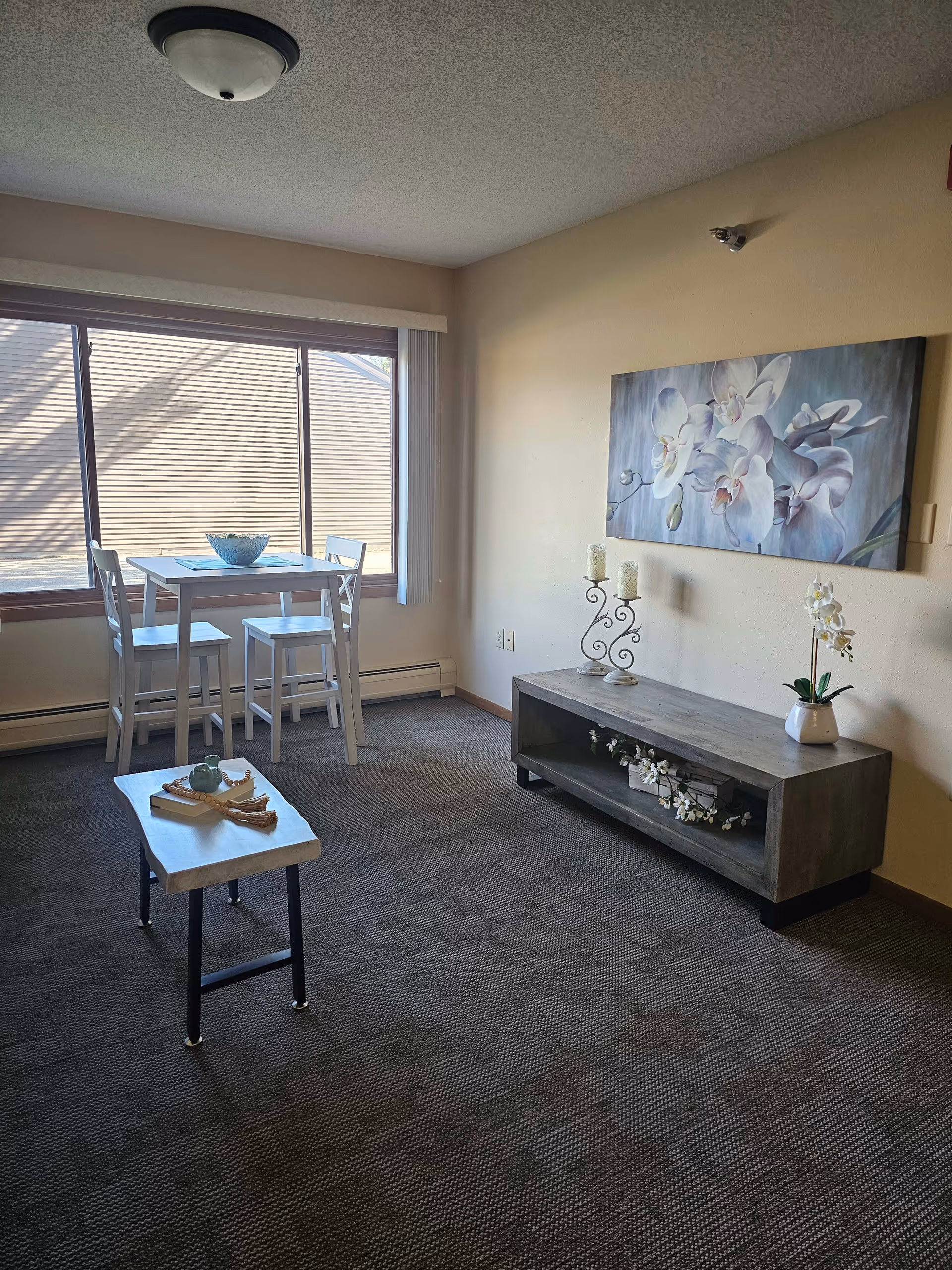 A small living area with a window letting in natural light. There is a tall white table with two matching chairs near the window. A low wooden console table against the wall holds decorative items including candles and a potted orchid. Above the console is a large painting of white orchids. In the foreground, a small wooden bench has a book and decorative items on it. The room has beige walls and a carpeted floor.
