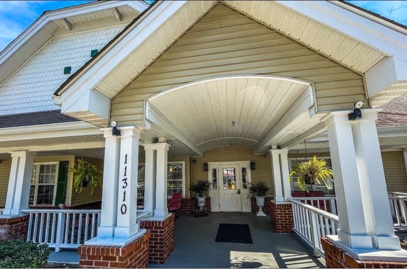 Entrance of a senior living facility with a covered porch supported by white columns on brick bases. The building has beige siding and white trim, with the number 11310 displayed on one of the columns. There are potted plants and seating visible near the entrance door.