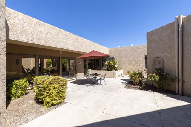 Sunny enclosed courtyard with a patio table, umbrella and chairs, surrounded by stucco walls and small shrubs.