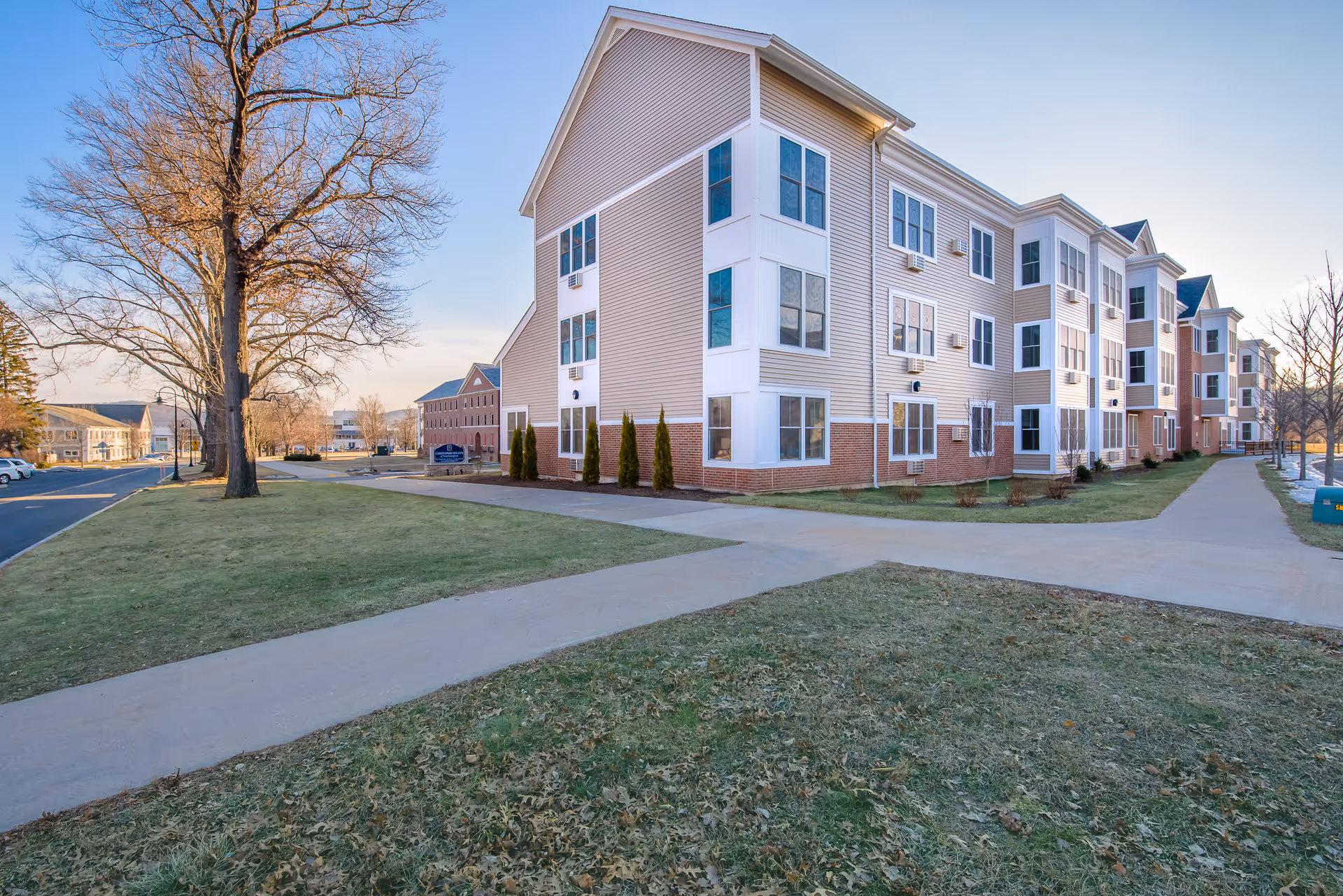 Exterior view of a multi-story senior living facility building with beige siding and white trim, surrounded by sidewalks and grassy areas with some trees, under a clear sky.