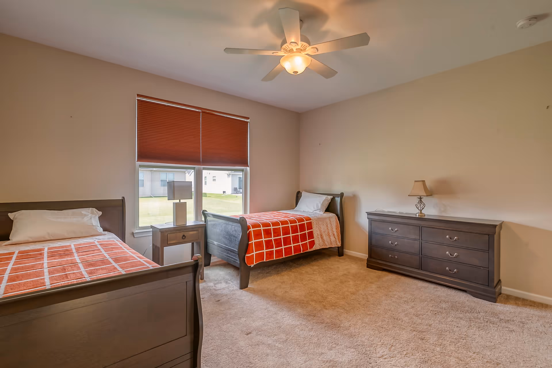 A bright bedroom with two single beds, a dresser, nightstand, ceiling fan and a window with red blinds.