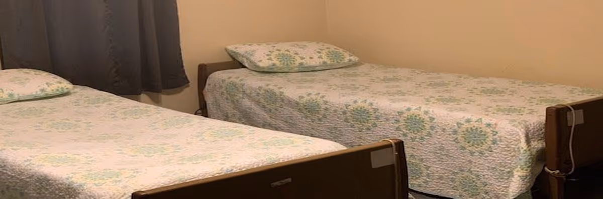 A simple bedroom with two single beds placed parallel to each other, each bed covered with a light-colored quilt featuring a green and blue floral pattern. There is a pillow on each bed with matching pillowcases. The room has beige walls and a window with dark curtains.