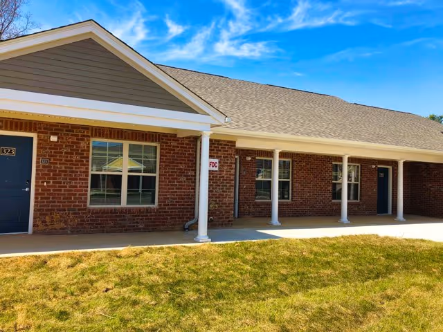 Exterior view of a single-story brick building with a covered walkway supported by white columns. The building has multiple windows and doors, with a grassy area in front under a blue sky with some clouds.
