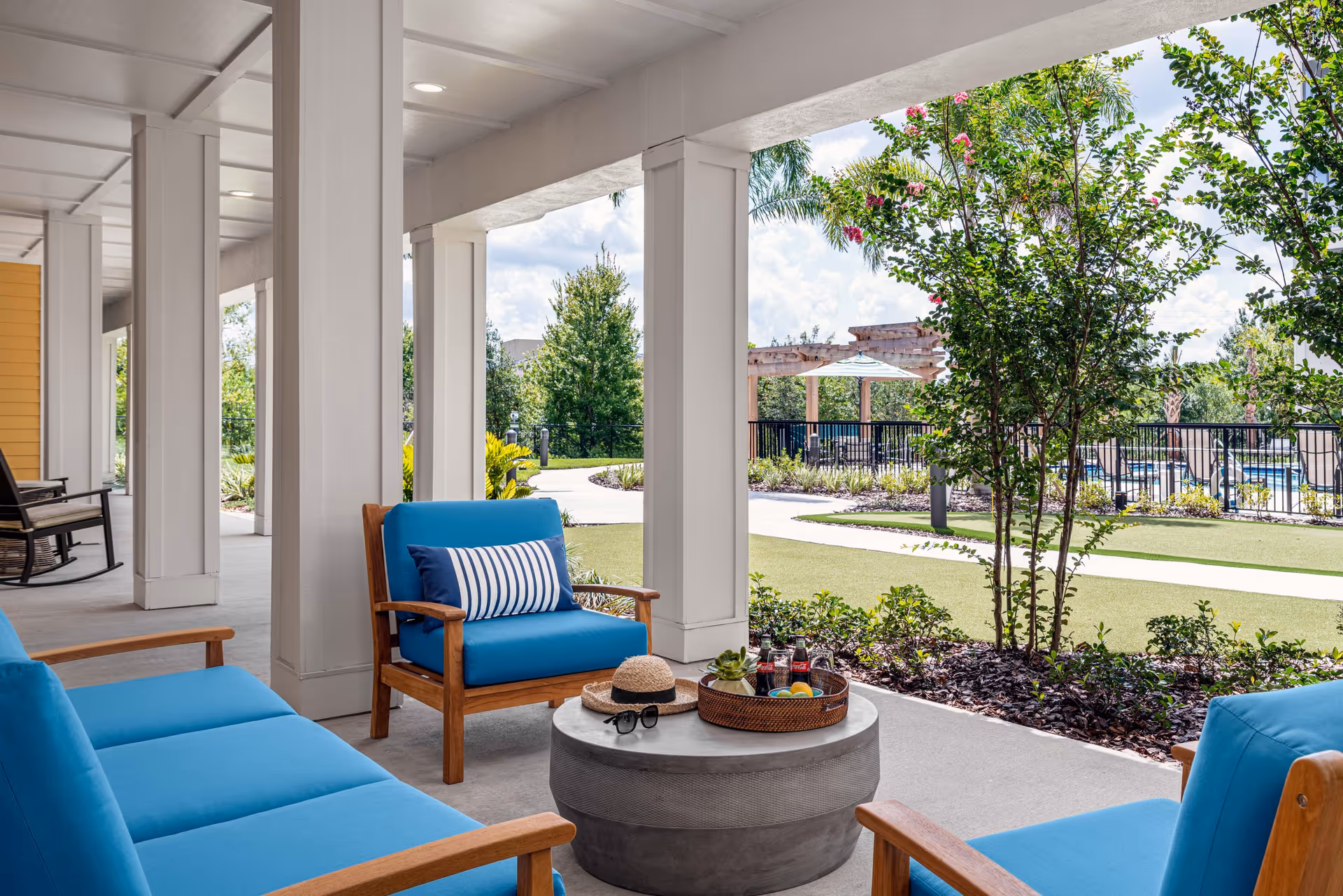 Outdoor covered patio area with blue cushioned wooden chairs and a matching sofa around a round concrete table. On the table are a straw hat, sunglasses, a small plant, and a tray with drinks and fruit. The patio overlooks a landscaped garden with trees, shrubs, a walking path, and a pergola near a fenced pool area under a partly cloudy sky.