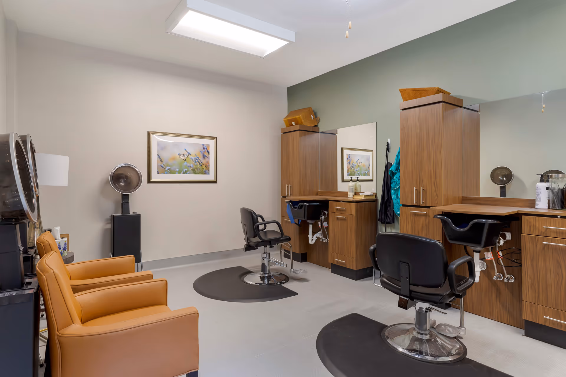 Interior view of a senior living facility's hair salon with two black salon chairs in front of wooden cabinets and sinks, two tan waiting chairs, a hair dryer, and a framed picture of flowers on the wall.