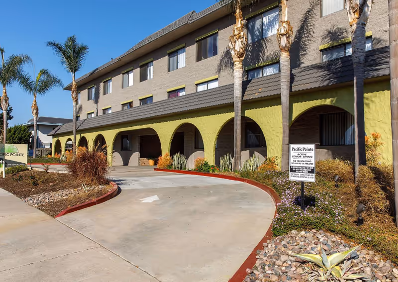 Exterior view of Pacific Pointe Senior Apartments with a curved driveway, arches, and palm trees.