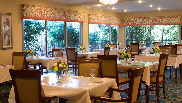 Bright dining room with multiple tables set with white tablecloths, chairs, floral centerpieces and large windows overlooking greenery.