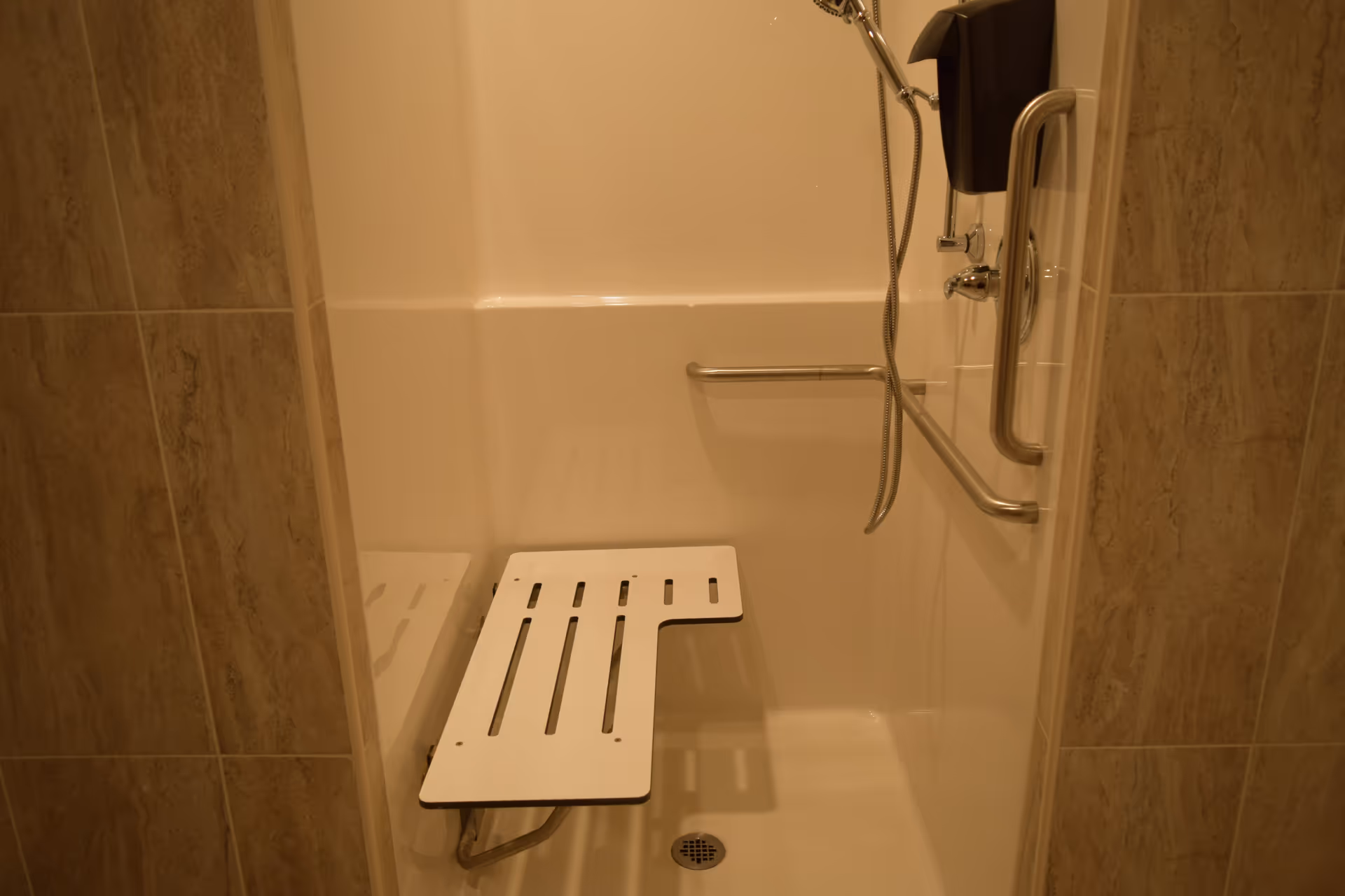 A shower area with beige tiled walls, a white fold-down shower seat, a handheld showerhead, and a horizontal grab bar for support.